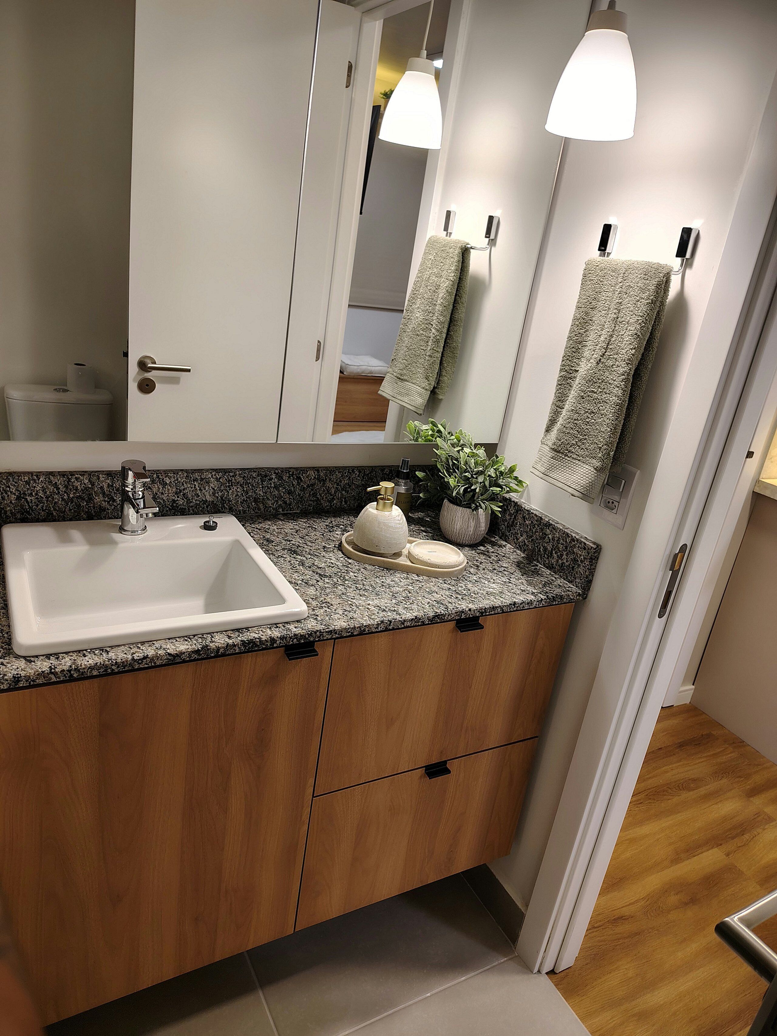 Un baño moderno con encimera de granito, un lavabo blanco, armarios de madera, un espejo y dos luces empotradas.