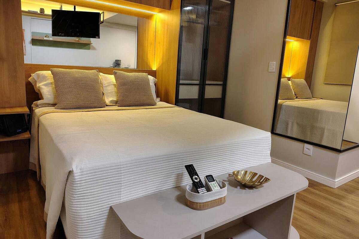 Une chambre d’hôtel comprenant un lit, un téléviseur fixé au mur, une table de chevet avec une télécommande, un vase décoratif et un miroir.