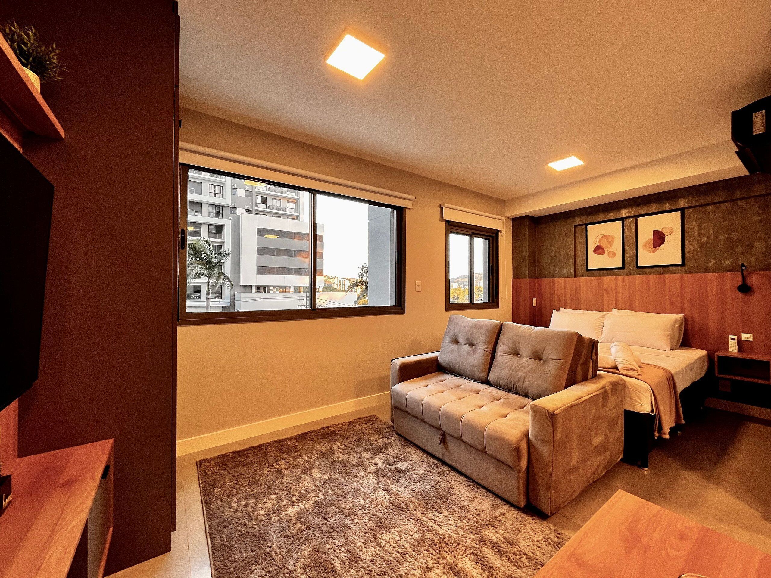 A hotel room with a sofa, a bed, a wooden headboard, and a view of buildings outside the window.