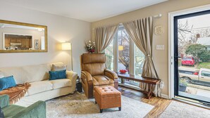 Un salon comprenant un canapé, un fauteuil, un pouf et une grande fenêtre avec des rideaux.