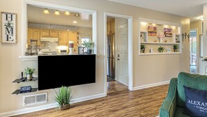Un salon moderne avec un téléviseur à écran plat, un fauteuil vert et une cuisine visible à travers une porte ouverte.