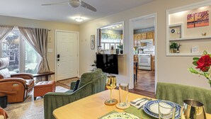 Un coin repas avec une table dressée pour deux personnes, un téléviseur en arrière-plan et une cuisine visible à travers une porte ouverte.