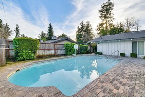A rectangular swimming pool with a brick deck, surrounded by a wooden fence and greenery.
