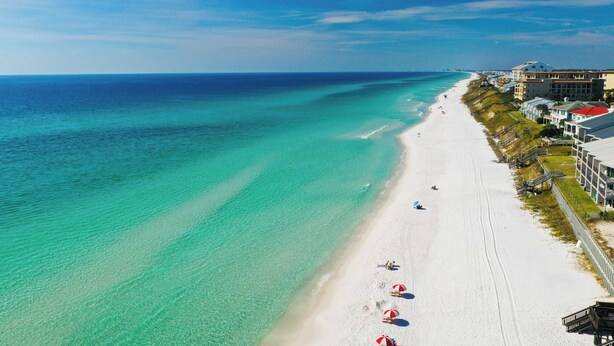 Ein Strand mit klarem türkisblauem Wasser, weißem Sand und einer Reihe von Gebäuden mit Balkonen entlang der Küste.