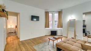 Een moderne woonkamer met een houten vloer, een beige bank, een flatscreen televisie, een grote spiegel en een radiator bij een raam met beige gordijnen.