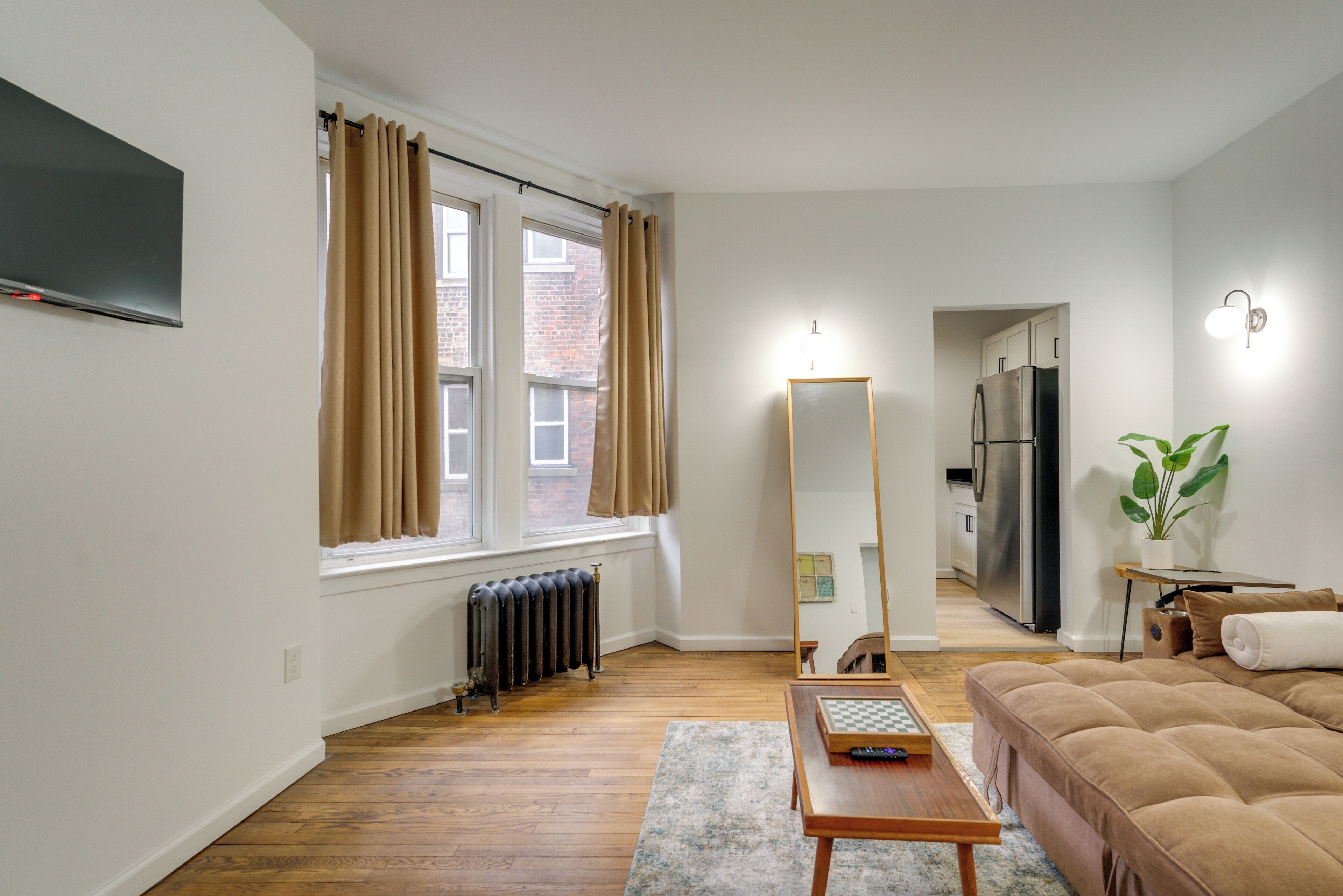 Un salon avec un canapé, une table, un miroir, un radiateur, un téléviseur et une fenêtre avec des rideaux.