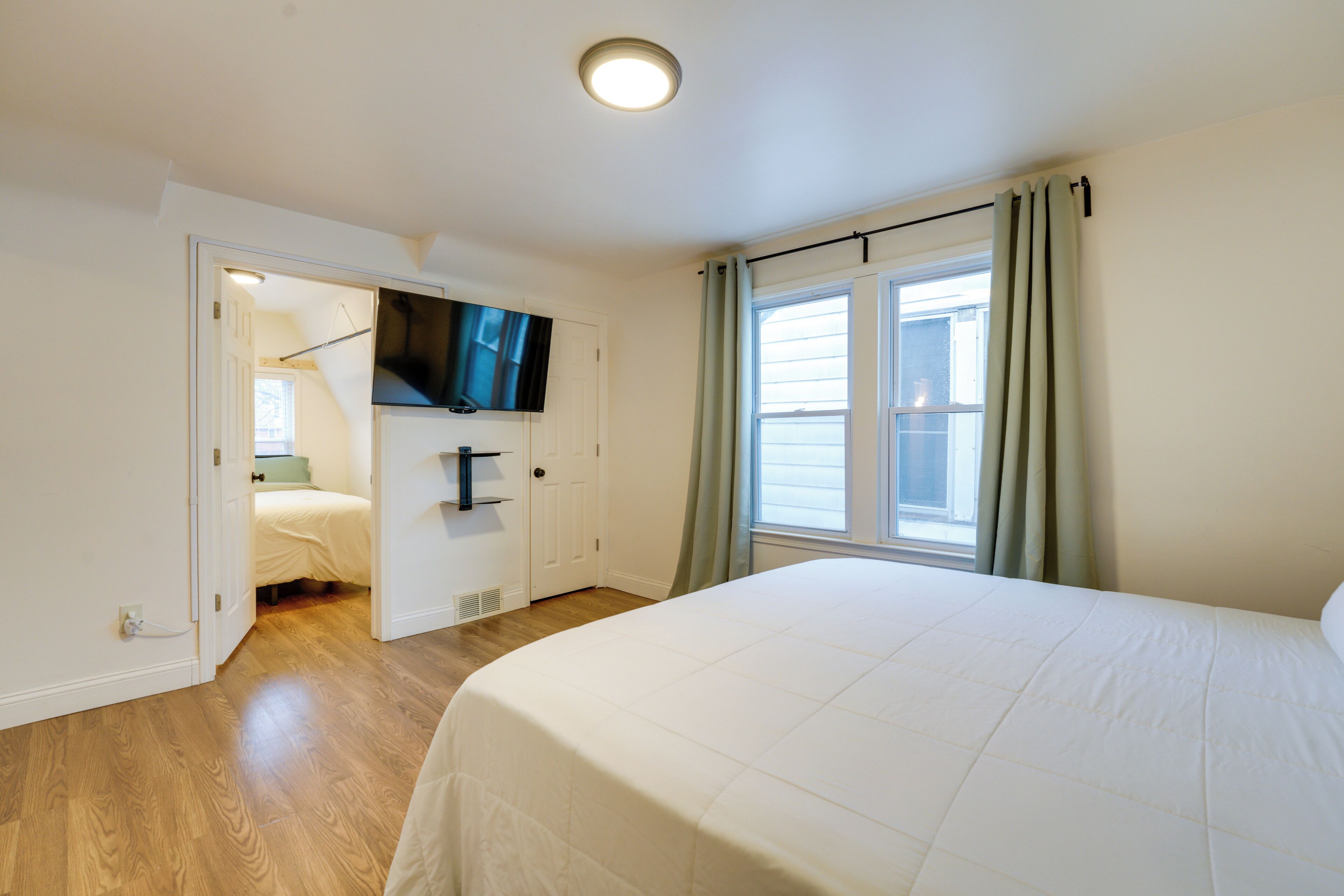 Una camera da letto con un letto, una televisione fissata al muro, una finestra con tende e una porta che dà su un'altra stanza.