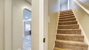 Un escalier recouvert de moquette, doté d’une rampe, mène à une pièce avec une porte blanche et une grille d’aération sur le mur.