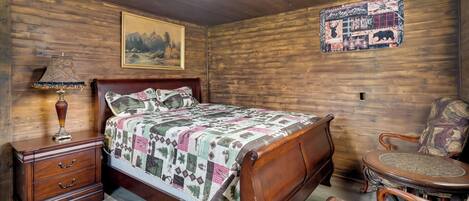 Un dormitorio de madera con cama, mesita de noche, lámpara, silla, alfombra y un cuadro y un póster en la pared.