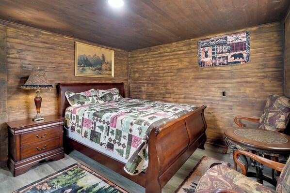 Un dormitorio de madera con cama, mesita de noche, lámpara, silla, alfombra y un cuadro y un póster en la pared.