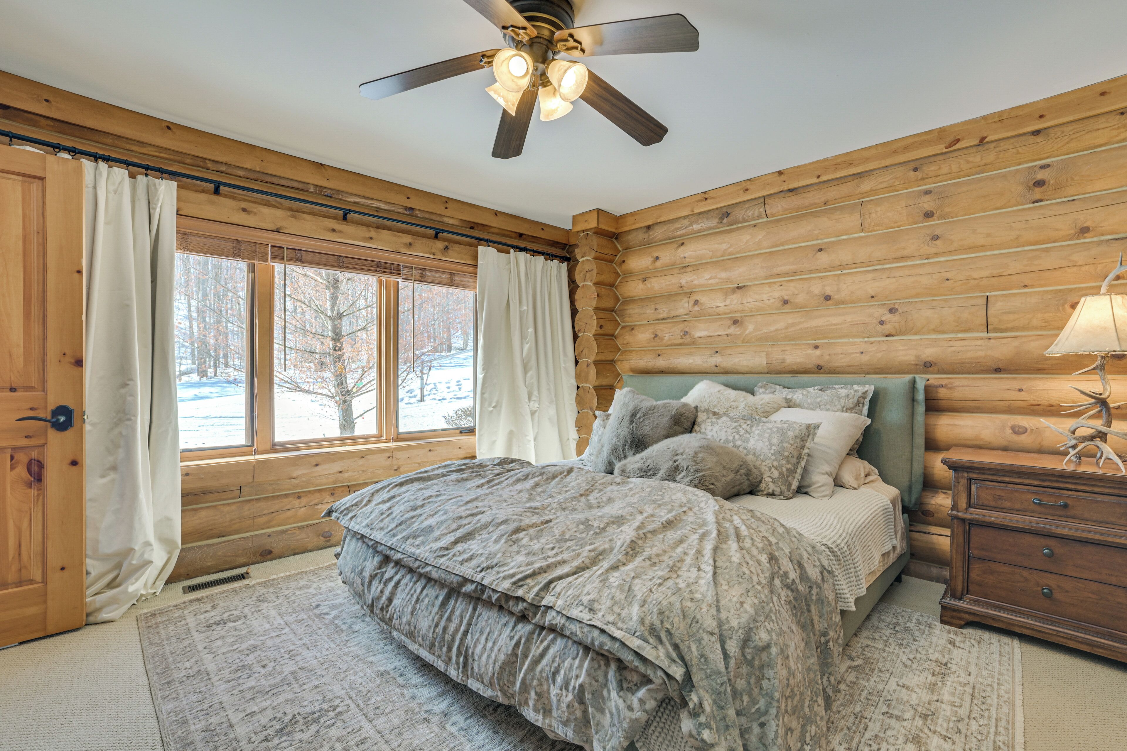 Un dormitorio de cabaña de madera con cama, mesita de noche, ventilador de techo y ventana con cortinas.
