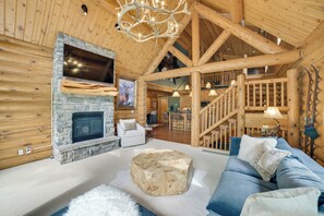 A log cabin interior with a stone fireplace, a large TV mounted above it, a wooden staircase, and a cozy seating area with a blue sofa and white cushions. - Ski-in, Ski-out Group Getaway in Harbor Springs! (Harbor Springs)