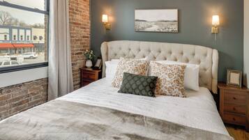 A bedroom with a tufted headboard, a large window with curtains, a view of a street, and two wall-mounted lamps.