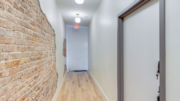 Um corredor com parede de tijolos, piso de madeira, porta branca e luminária no teto.