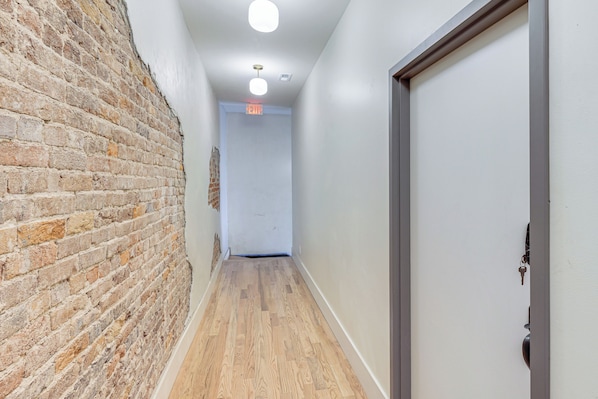A hallway with a brick wall, a wooden floor, a white door, and a ceiling light fixture.