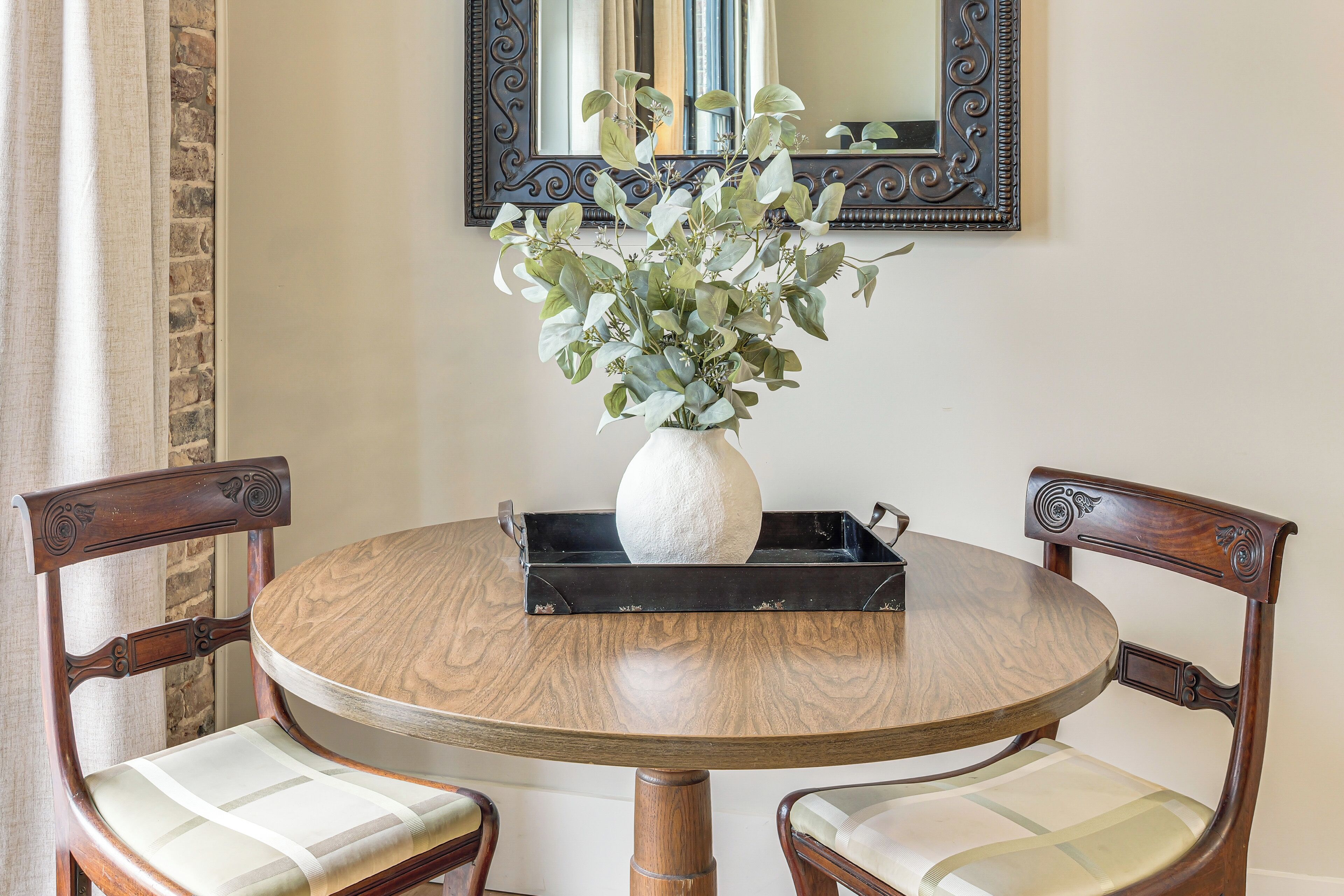 A round wooden table with a white vase and green foliage centerpiece, flanked by two upholstered chairs with wooden frames.