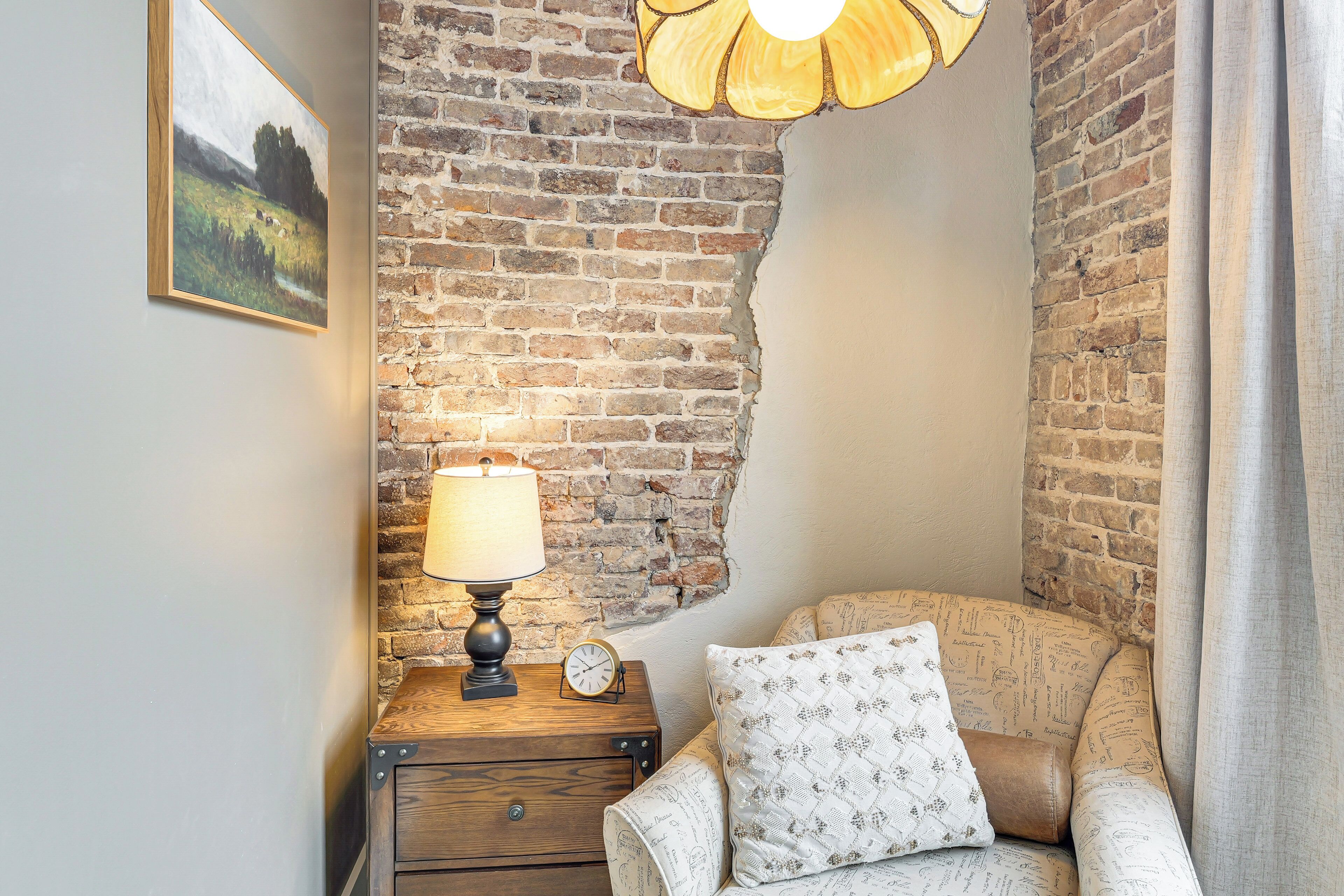 Una camera accogliente con un muro in mattoni, un comodino in legno con una lampada, una sveglia e un divano decorato.
