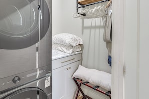 A compact laundry area with a washing machine, a drying rack, and a small bed with pillows.