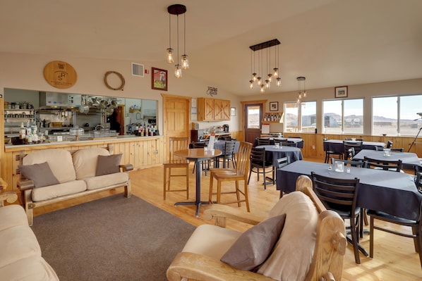 A spacious dining area with wooden tables and chairs, a kitchen in the background, and large windows offering a view.
