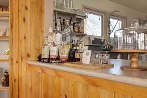 A bar area with wooden shelves, various liquor bottles, glasses, and a large glass container on a wooden stand.
