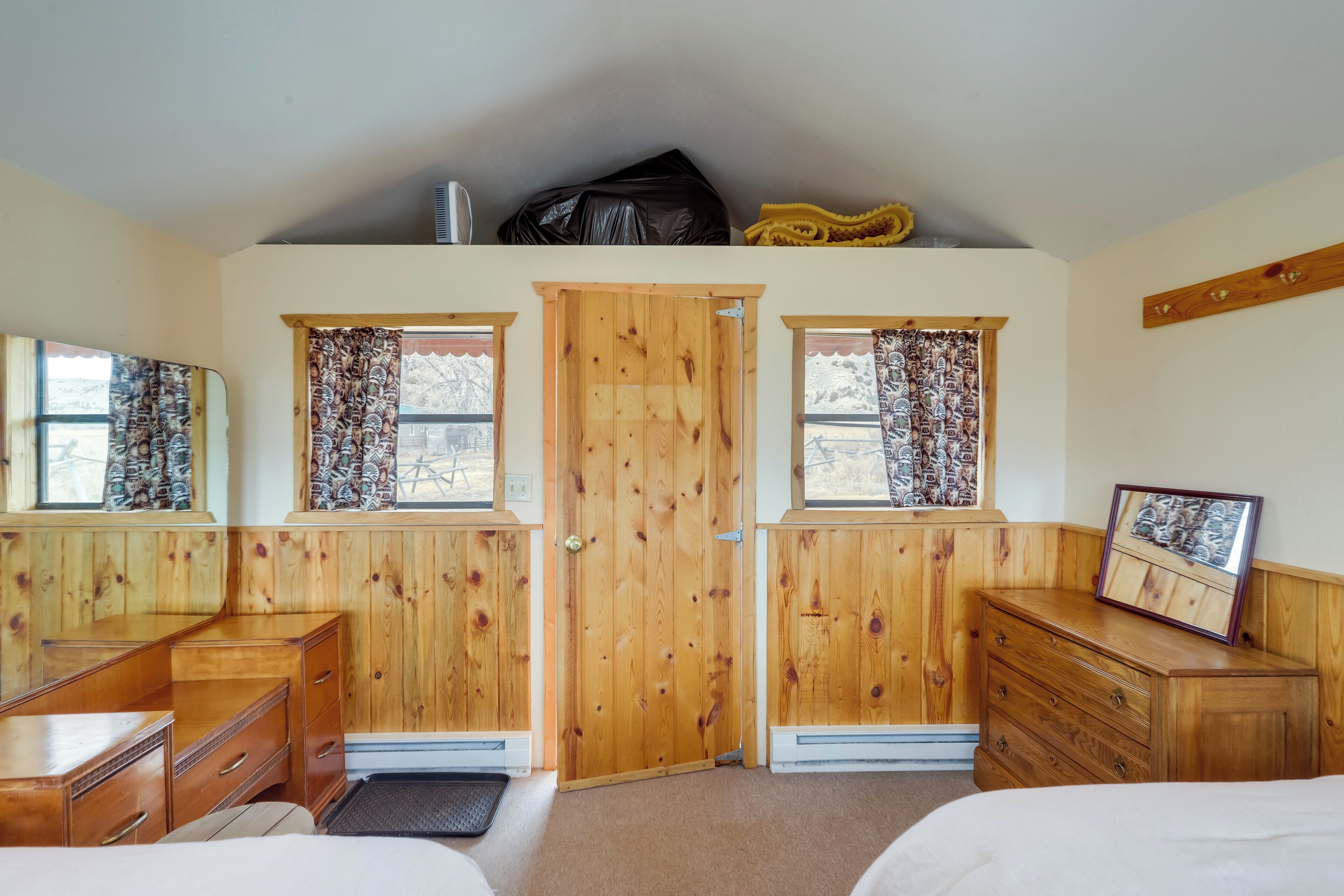 A room with wooden walls, a central door, two windows with patterned curtains, a bed, a dresser, and a framed picture on the dresser.