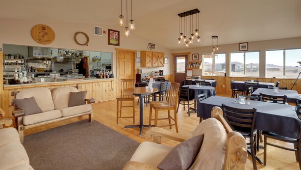 A spacious dining area with wooden tables and chairs, a kitchen in the background, and large windows offering a view.