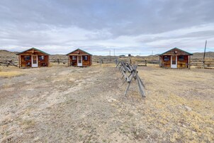 Tres cabañas de madera con suelo de tierra y una cerca de madera en un entorno rural.