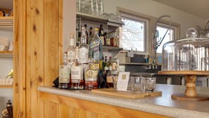 A bar area with wooden shelves, various liquor bottles, glasses, and a large glass container on a wooden stand.