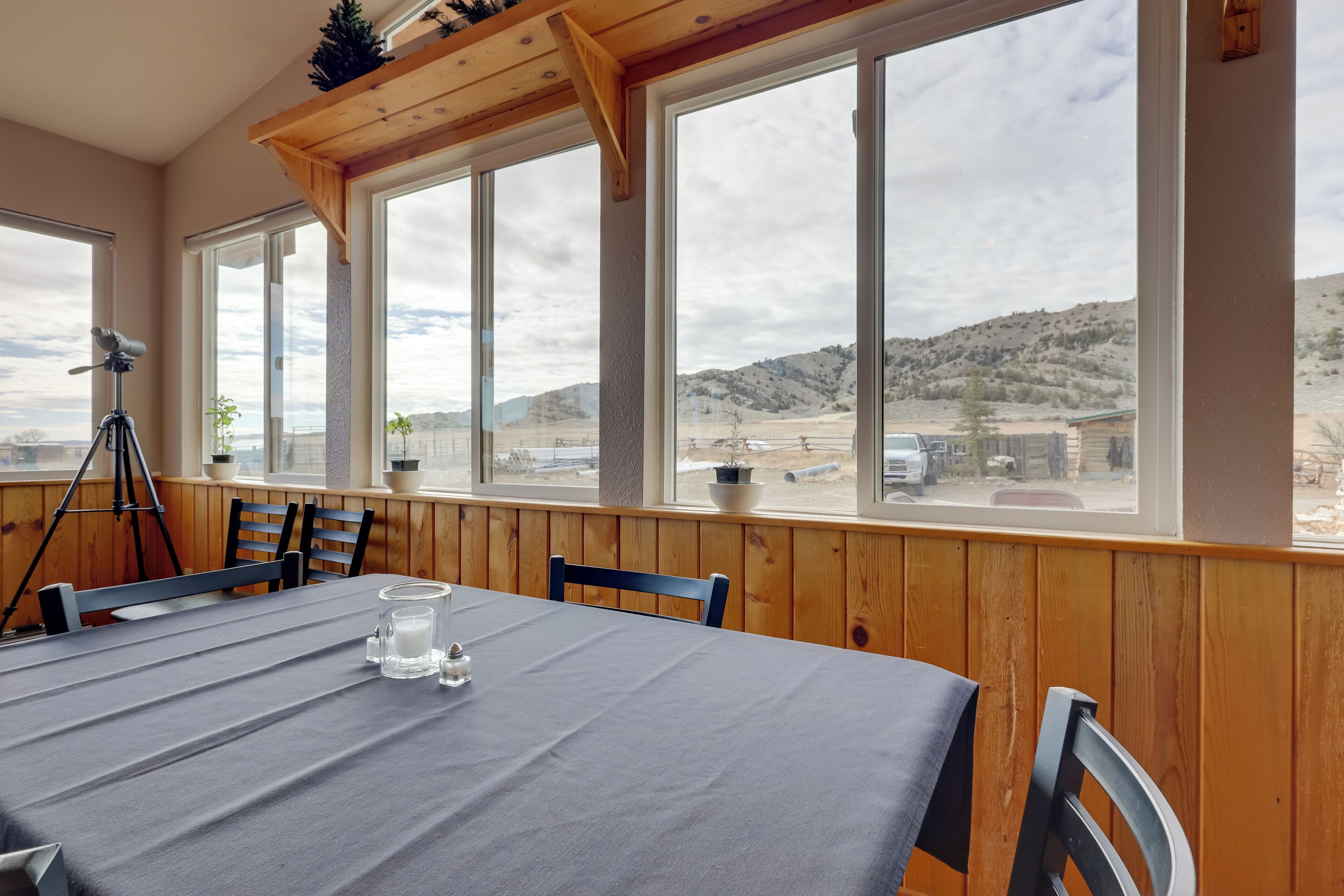Un coin repas avec une table dressée pour deux personnes, de grandes fenêtres offrant une vue sur un paysage désertique, des murs lambrés et un trépied dans un coin.