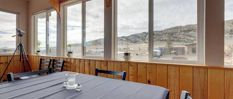 Un coin repas avec une table dressée pour deux personnes, de grandes fenêtres offrant une vue sur un paysage désertique, des murs lambrés et un trépied dans un coin.