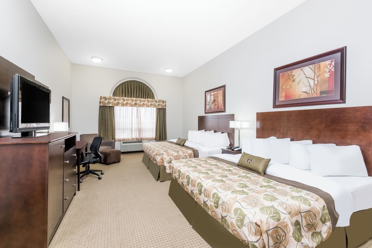 A hotel room with a TV, desk, two beds, a chair, a desk lamp, a framed picture, and a window with curtains.