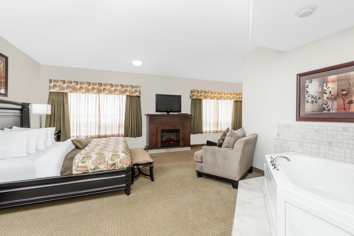 A hotel room with a bed, a sofa, a chair, a fireplace, a television, and a bathtub.
