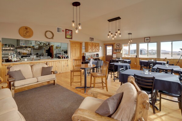 A spacious dining area with wooden tables and chairs, a kitchen in the background, and large windows offering a view.