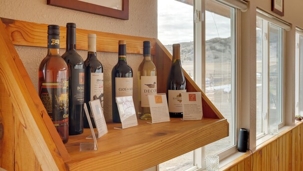 Exhibición de vinos con botellas y etiquetas, un estante de madera y una ventana con vistas.