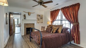 Un dormitorio con cama, ventilador de techo, chimenea y ventana con cortinas.