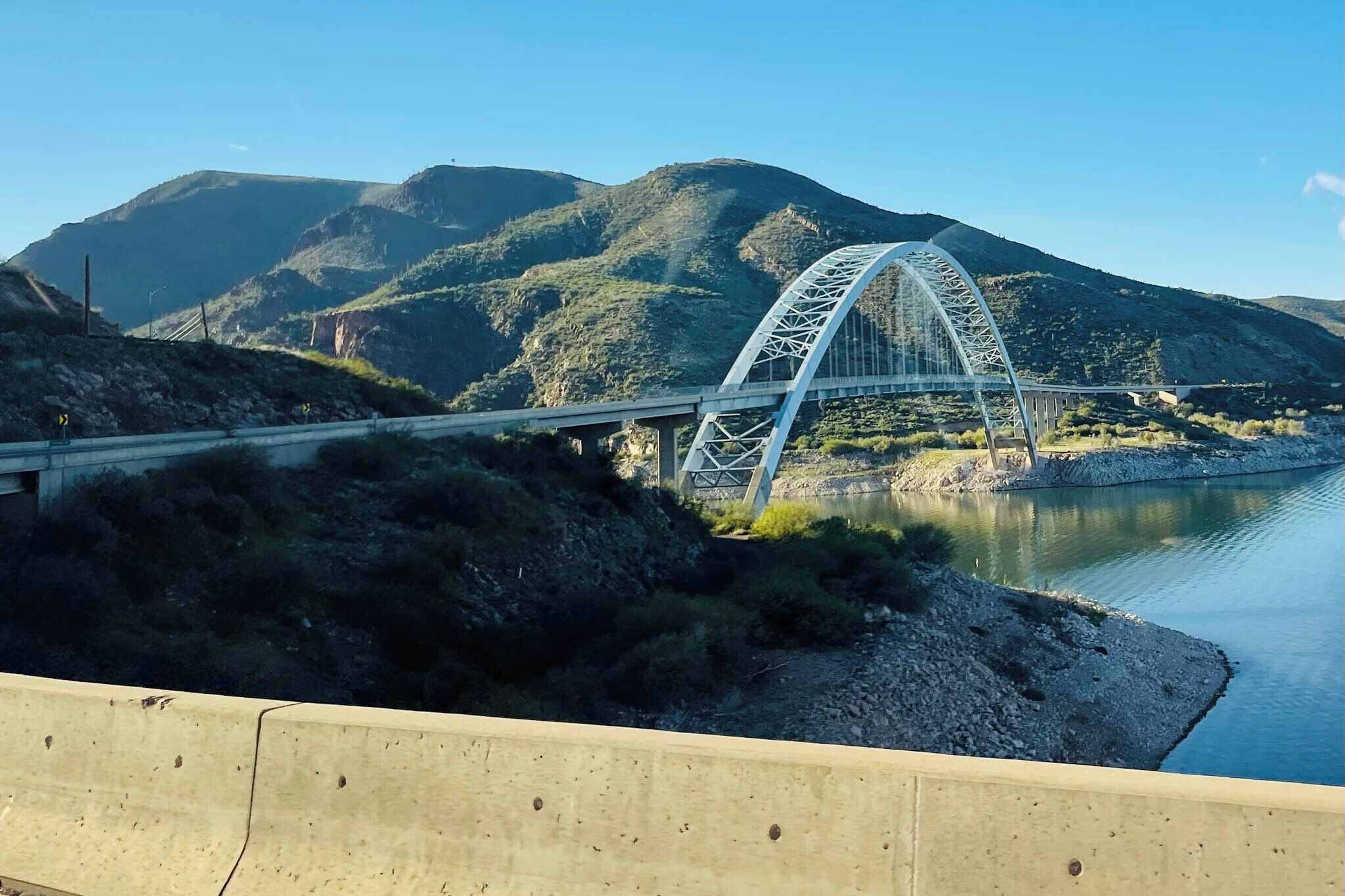 A bridge with a steel arch design over a river, surrounded by hills and vegetation.