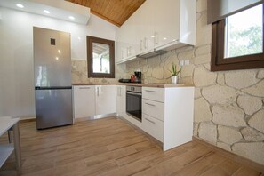 A modern kitchen with white cabinets, a stainless steel refrigerator, an oven, and a stone wall. - Ikarion Village Home by RentalsPro (Skala Fourkas)