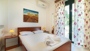 A hotel room with a bed, two bedside tables with lamps, a painting on the wall, and a window with floral curtains. - Aegean Maisonette by RentalsPro - Kryopigi (Kriopighi)