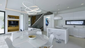 A modern kitchen with white cabinetry, a central island, and a dining area with a glass table and white chairs. - Μaraviglia Ethereon Villa by RentalsPro (Nikiti)