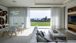 A modern living room with a large window, a sofa, a coffee table, and a TV. - Μaraviglia Ethereon Villa by RentalsPro (Nikiti)