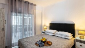A bedroom with a bed, a nightstand, a lamp, a window with curtains, and a view of the outside. - GnG Apartment 1 by RentalsPro - Nea Flogita (Nea Flogita)