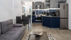 A modern kitchen with built-in appliances, a dining area with a gray sofa and chairs, and a small island. - GnG Apartment 1 by RentalsPro - Nea Flogita (Nea Flogita)