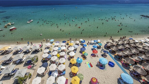 Una spiaggia con molti ombrelloni e lettini, un mare cristallino e imbarcazioni all'orizzonte.