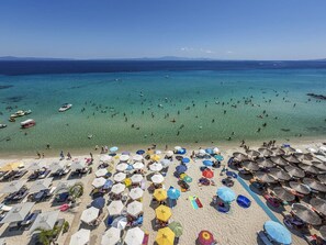 Una playa con numerosos sombrillas y tumbonas, un mar azul cristalino y barcos a lo lejos.