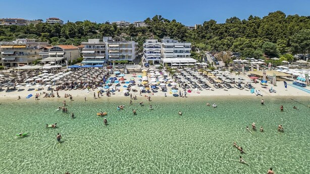 Ein Strand mit klarem Wasser, Sonnenschirmen und Gebäuden im Hintergrund.