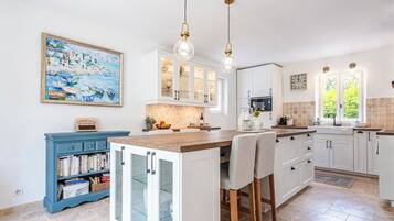 A kitchen with white cabinets, a blue bookshelf, a large island with chairs, pendant lights, and a painting on the wall.