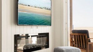 Ein modernes Wohnzimmer mit einem flachbildfernseher an einer weiß vertikalen Wand, einem eingebauten Kamin, einem runden Couchtisch und Blick auf den Strand durch ein großes Fenster.