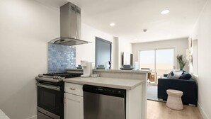 Eine moderne Küche mit Herd, Backofen und Geschirrspüler, einer blauen Küchenzeile und Blick in einen Wohnbereich mit Sofa und Pflanze.