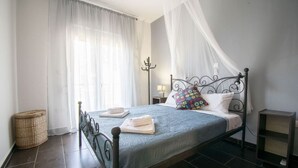 A neatly made bed with a blue bedspread, white pillows, and a colorful floral pillow. There is a bedside table with a lamp, a wicker basket, and a small shelf. The room has a dark tiled floor and white walls. - Sunlight  Maisonette by RentalsPro (Nikiti)
