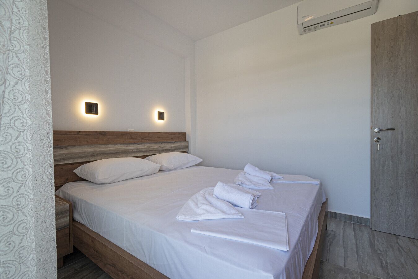 Une chambre à coucher avec une tête de lit en bois, deux lampes de chevet, un lit avec du linge de lit blanc et une porte sur le côté droit.